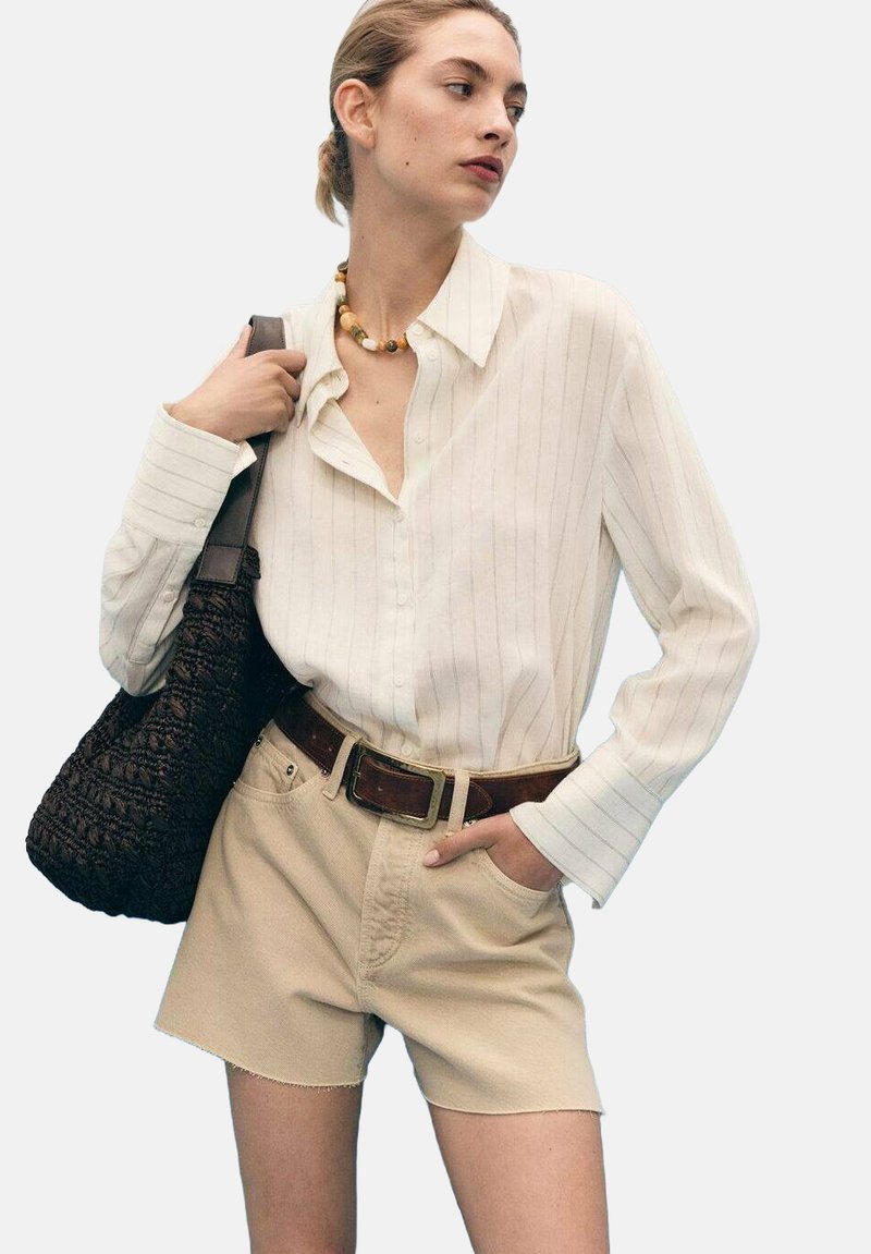 Chemise boutonnée à rayures blanches, shorts beiges, ceinture marron et sac marron texturé. Le modèle porte un collier en perles volumineux. Palette de couleurs neutres.