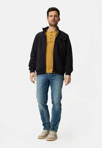 Schwarze Fleecejacke mit Reißverschluss über einem gelb gestreiften Henley-Shirt, kombiniert mit hellblauen Jeans und beigen Sneakers. Stehend vor weißem Hintergrund.