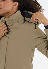 Lysbrun softshell-jakke med høj krave, justerbar snøre og sorte detaljer. Struktureret stof med synlige sømme og knapper.