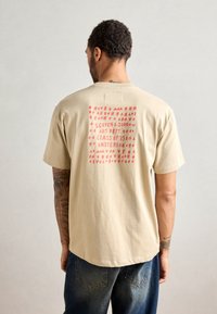 Beige színű pamut póló, amelyen piros nyomott grafika található geometriai formák és a "SCOTCH & SODA ART DEPT. CLASS OF '85 AMSTERDAM" felirattal a hátsó részen.