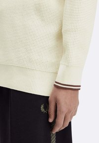 Maglione color crema con un motivo a maglia texture tipo waffle e polsini a coste, caratterizzati da due strisce marroni. Indossato con pantaloni neri.