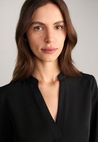 Czarna bluzka z głębokim dekoltem w kształcie V, minimalnym kołnierzykiem i gładką teksturą, prezentująca elegancki design. Prosta i elegancka, bez ozdób.
