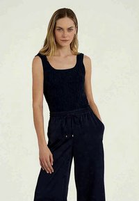 Jovem mulher com cabelo loiro liso, vestindo uma blusa preta de malha sem mangas e calças largas pretas, com as mãos nos bolsos.