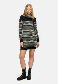 Strickkleid in Schwarz und Creme mit geometrischen Mustern, Kragen, langen Ärmeln und gerippten Bündchen. Kombiniert mit schwarzen Stiefeln.