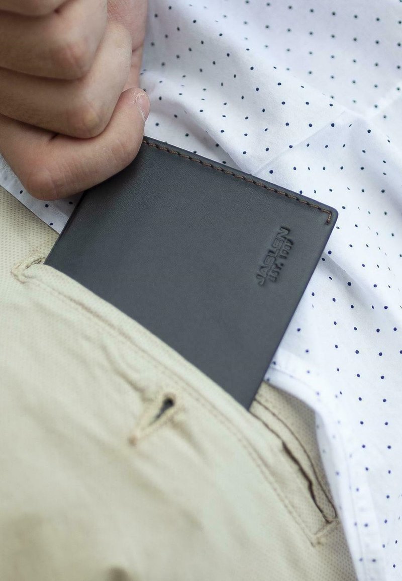 Billetera de cuero negro parcialmente visible en un bolsillo, con costuras precisas y un logo en relieve. Camisa blanca con pequeños puntos azules en el fondo.
