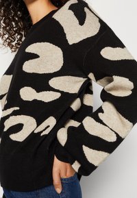 Schwarzer Pullover mit beigem abstraktem Muster, weicher Textur, langen Ärmeln und geripptem Saum. Die Hand des Models befindet sich vorne, es trägt eine blaue Jeans.