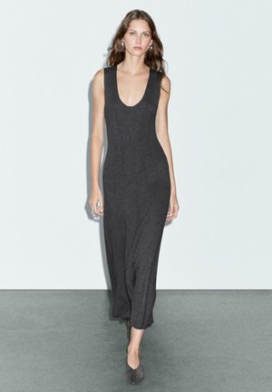 Donna che cammina in un vestito midi aderente senza maniche di un grigio scuro con scollatura profonda e scarpe a punta coordinate su sfondo neutro.