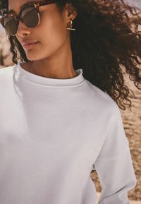 Weißes Sweatshirt mit lockerer Passform und Rundhalsausschnitt. Das Modell trägt Schildpatt-Sonnenbrillen und goldene Ohrringe mit einem horizontalen Balken-Detail.