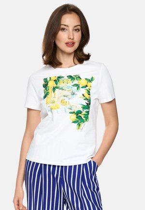 Woman wearing white short-sleeve shirt with lemon and coastal landscape print, paired with blue and white vertical striped pants.