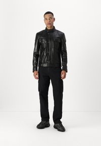 Veste en cuir noir avec un col montant, des poches à bouton et une coupe ajustée. Associée à un pantalon cargo noir et des baskets texturées.