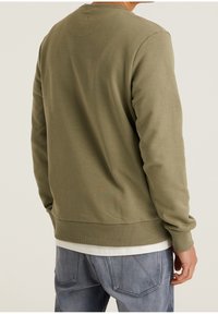 Sudadera de cuello redondo en verde oliva con puños y dobladillo de canalé, hecha de una tela suave, que presenta un reverso texturizado y una camisa blanca en capas por debajo.