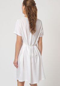 Vestido blanco de manga corta con diseño de lazo en la espalda, hecho de tela ligera. Presenta un ajuste holgado y discretos bolsillos laterales.