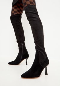 Botas altas de ante negro con punteras afiladas, tacones altos y delgados, y cremalleras laterales; textura suave y diseño ajustado.