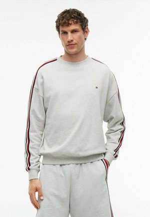 Mann mit lockigem Haar trägt hellgrauen Pullover und passende Hose mit roten, weißen und blauen Streifen an Ärmeln und Hosenbeinen.