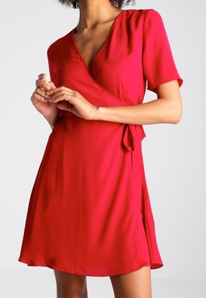 Kvinde iført en rød slå-om kjole med korte ærmer og knælængde, bundet i taljen, stående mod en ensfarvet lys baggrund.