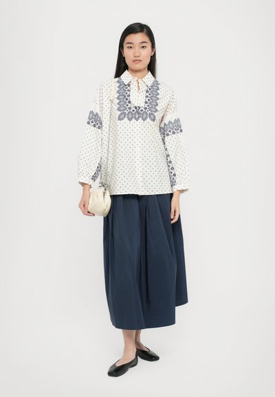 Weiße Bluse mit blauen, gemusterten Akzenten und Punkten, kombiniert mit einem knielangen, plissierten Rock in Navy-Blau und schwarzen Ballerinas. Hält eine kleine cremefarbene Tasche.