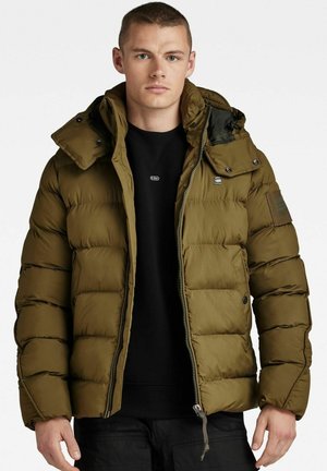 Chaqueta acolchada verde oliva con cierre de cremallera y botón, capucha desmontable y puños acanalados. Presenta un tejido acolchado y un parche en la manga.