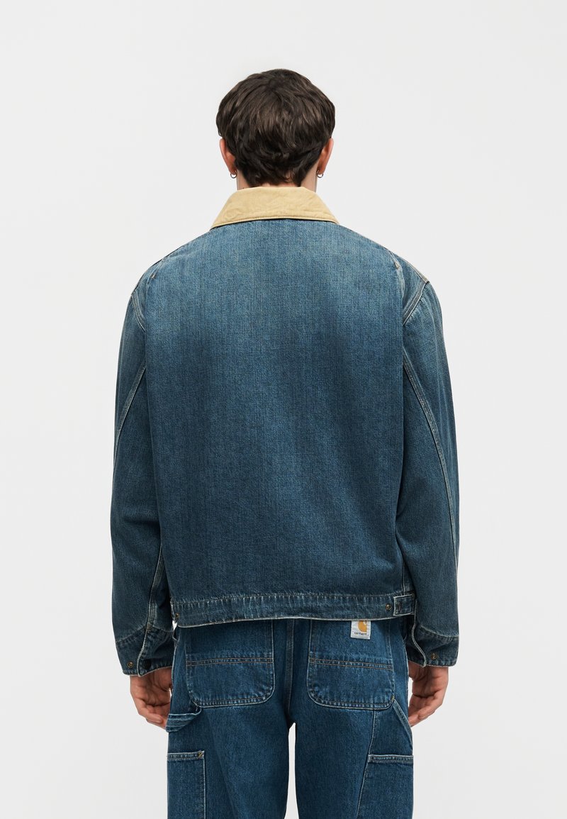 Persona vista da dietro che indossa una giacca di jeans blu con colletto in velluto beige e pantaloni di jeans blu abbinati con tasche e toppa con etichetta.