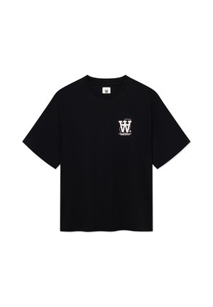 Ung person med langt rødt hår iført en sort grafisk t-shirt, brune løse bukser og sorte sko, stående med hænderne i lommerne.