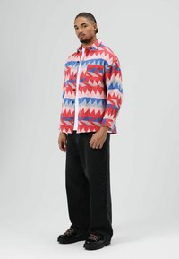 Bunt gemustertes Hemd mit roten, blauen und weißen dreieckigen Designs. Das Material wirkt strukturiert. Kombiniert mit schwarzen Weithose und Stiefeln.