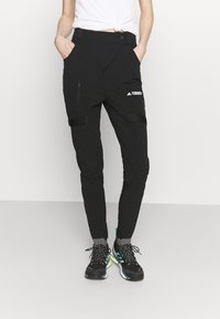 Pantalones exteriores negros con bolsillos con cremallera, correas ajustables y un diseño ajustado. Fabricados con material duradero, combinados con zapatos negros y verdes.
