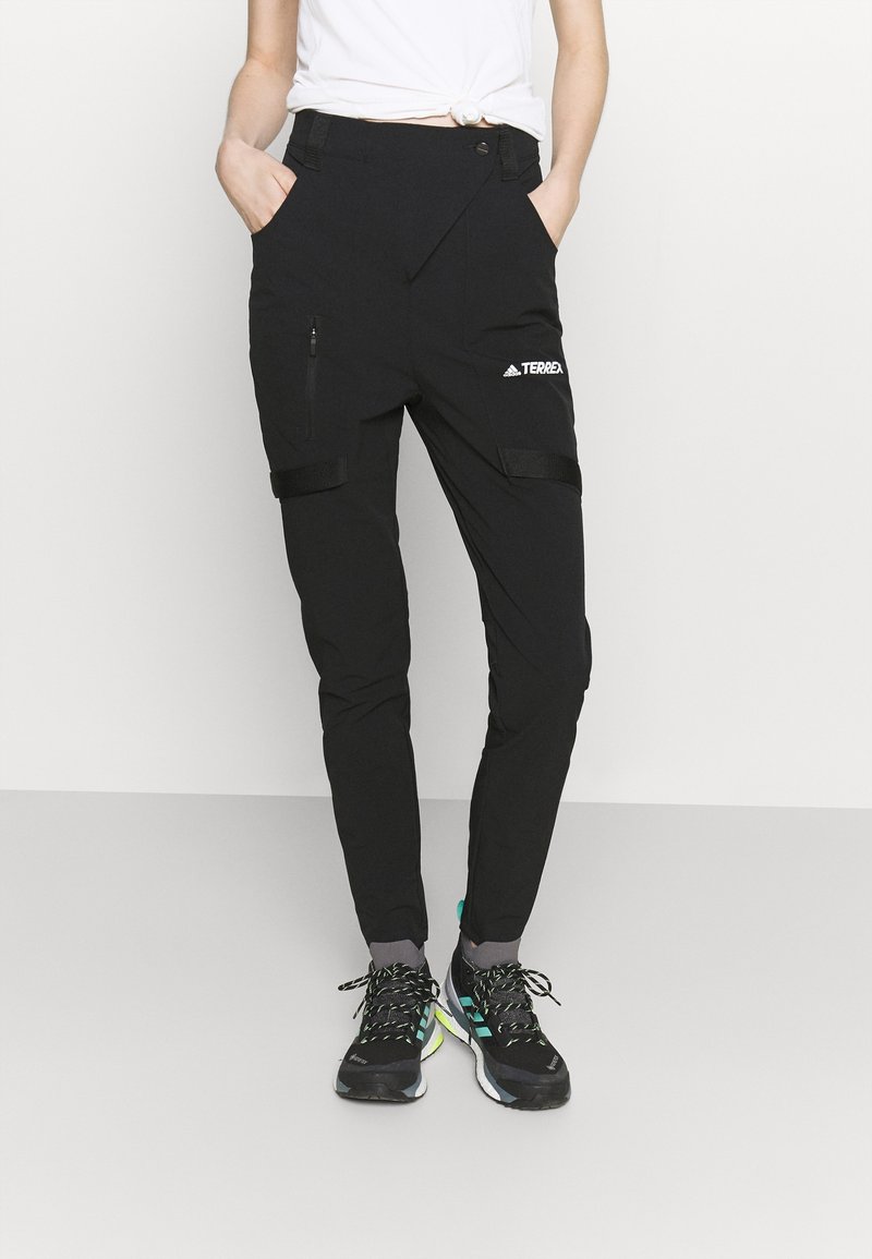 Pantalones exteriores negros con bolsillos con cremallera, correas ajustables y un diseño ajustado. Fabricados con material duradero, combinados con zapatos negros y verdes.