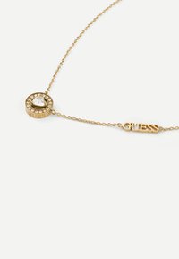 Collana in tono dorato con un pendente circolare adornato da una pietra trasparente circondata da pietre più piccole, e un accento con il logo "GUESS".
