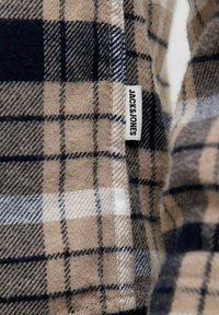 Beżowa, czarna i biała tkanina w kratkę z metką odzieżową "JACK & JONES" wszytą w szew.