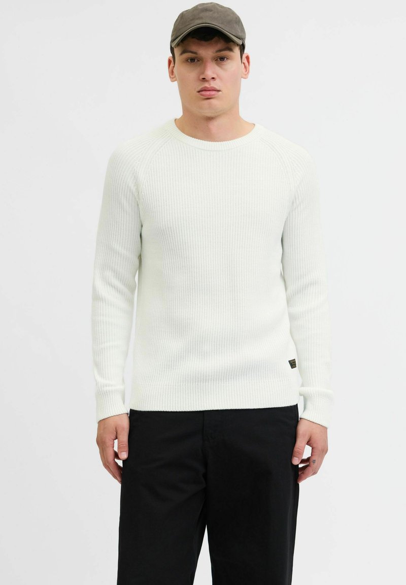 Maglione bianco a costine con collo rotondo e maniche lunghe, caratterizzato da una vestibilità attillata e dettagli con etichetta nera, abbinato a pantaloni neri.