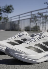 Weiße Sportschuhe mit drei schwarzen Streifen, strukturiertem Mesh-Obermaterial, gepolsterter Sohle und verstärkter Zehenkappe, auf einer flachen Oberfläche platziert.