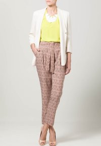 Bílý blazer přes jasně žlutý top, doplněný o vzorované, vysoké béžové kalhoty. Modelka má na sobě nahé vysoké podpatky a výrazný náhrdelník.