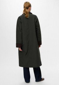 Cappotto lungo imbottito verde con colletto nero, polsini marroni e una texture liscia. Presenta una linea dritta e si estende al di sotto del ginocchio.