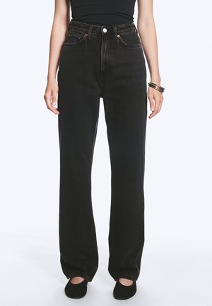 Femme portant un jean noir taille haute à jambes larges, un haut noir, des chaussures noires et des bracelets à un poignet, debout devant un fond uni.