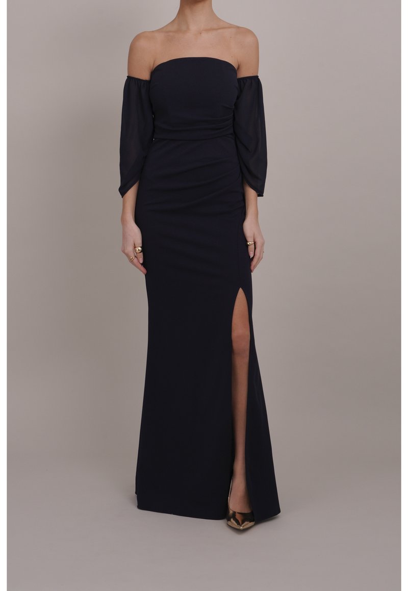 Femme portant une robe de soirée noire épaules dénudées avec des manches longues transparentes et une fente haute, debout devant un fond uni.