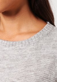 Ljust grå stickad tröja med ribbad halsringning, som visar ett texturerat tyg och en lös passform, som betonar en enkel, minimalistisk design.