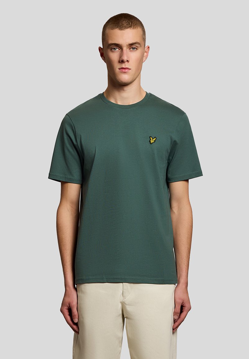 Νέος άνδρας με κοντά μαλλιά, που φοράει μια σκούρα πράσινη κοντομάνικη μπλούζα με μικρό κίτρινο λογότυπο πουλιά και ανοιχτό μπεζ παντελόνι.