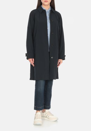 Cappotto impermeabile blu marina, due tasche frontali, chiusura a bottoni, con colletto. Indossato sopra una camicia azzurro chiaro e abbinato a jeans blu e sneaker.