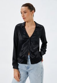 Zwarte satijnen blouse met een V-hals, lange mouwen en knoopsluiting. Gecombineerd met lichtblauwe versleten jeans. Gladde textuur, minimalistisch ontwerp.