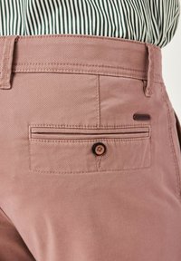 Helle rosa Baumwollhose mit einer Gesäßtasche, ausgestattet mit einem braunen Knopf und subtiler Textur, kombiniert mit einem grün-weiß gestreiften Hemd.