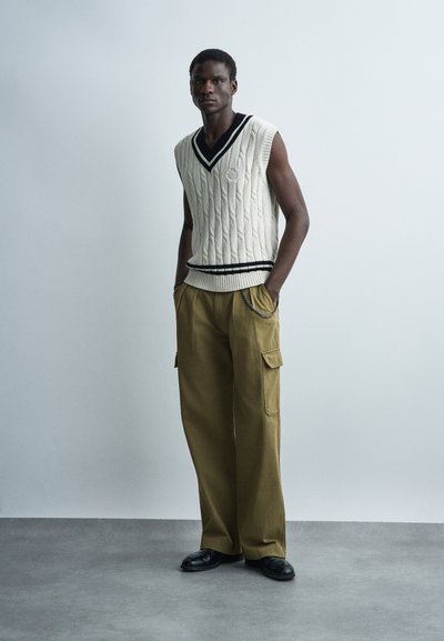 Άνδρας που φοράει αμάνικο εκρού πλεκτό γιλέκο με μαύρες ρίγες, φαρδιά χρυσαφί παντελόνια cargo και μαύρα παπούτσια, στέκεται μπροστά σε απλό φωτεινό φόντο.