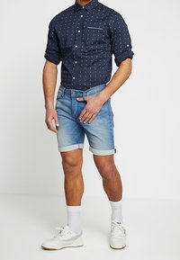 Granatowa koszula z guzikami z białymi kropkami, krótkie dżinsowe szorty i białe buty sportowe. Posiada podwinięte rękawy i kieszeń na piersi.