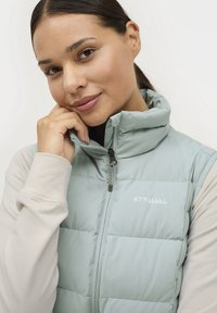 Lichtblauw puffervest met hoge kraag, ritssluiting en logo op de borst; gelaagd met een lichtbeige langslijfshemd. Gladde textuur.