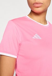 Ροζ αθλητική μπλούζα με λευκή επένδυση στον γιακά και λευκό λογότυπο Adidas. Η υφή του υφάσματος φαίνεται ελαφριά και διαπνέουσα.