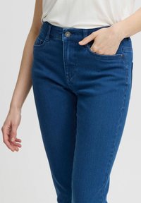 Tumedad teksased püksid, viie taskuga, eesnupu sulguriga ja kitsas lõikega. Materjal näib sile ja veidi veniv.