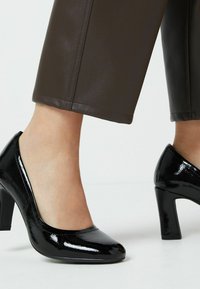 Black patent leather high heels with a rounded toe and a chunky heel. Worn with brown cropped pants, featuring a smooth, glossy texture.