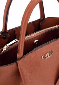 Borsa a tracolla in pelle marrone con texture, manici doppi, scomparto con zip e dettaglio del logo metallico. Presenta una chiusura a zip liscia.