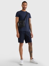 Mørkeblå t-skjorte og mørkeblå shorts kombinert med en svart crossover-veske. Vesken har teksturert materiale og glidelås, med logo detaljer. Hvite joggesko.