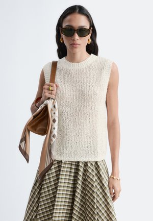 Femme portant des lunettes de soleil, un débardeur en maille, une jupe à carreaux, des bijoux dorés et tenant un sac marron avec un foulard à motifs noué à la poignée.
