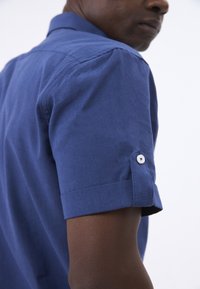 Camisa de manga corta de color azul marino con un detalle de botón para enrollar la manga. Textura de tela suave y corte ajustado. Costura de hombro invisible.