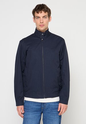 Mężczyzna w granatowej kurtce na zamek z wysokim kołnierzem i niebieskich dżinsach, stojący na jednolitym, jasnoszarym tle.